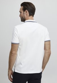 Hvit polo skjorte med ribbestrikket krage med marineblå striper. Korte ermer og splitt i kanten; bomullsteksturen er synlig.