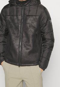 Svart jacka med quiltat övre avsnitt och slätt nedre parti. Har dragkedjestängning, huva och sidofickor. Mjuk textur, modern design.