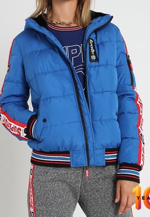 Femme portant une doudoune bleue avec des bandes rouges et blanches de marque sur les manches, un pantalon de survêtement gris avec des bandes assorties sur les côtés et un collier en chaîne dorée.