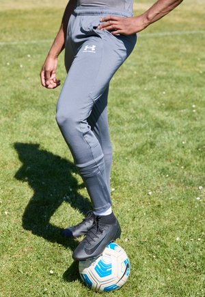 Persoon met grijze Under Armour broek en zwarte voetbalschoenen rust zijn voet op een wit-blauwe voetbal op een grasveld.
