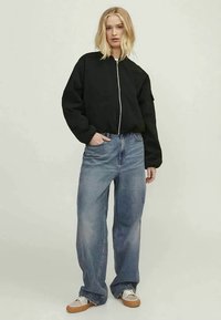Casaco bomber preto com fecho frontal, silhueta oversized e punhos canelados, combinado com jeans de ganga de perna larga em azul claro e sapatilhas bege.