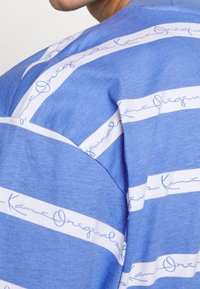 Blå kortärmad skjorta med vita ränder och kursiv text. Mjukt tyg med en avslappnad passform, sedd från sidan.