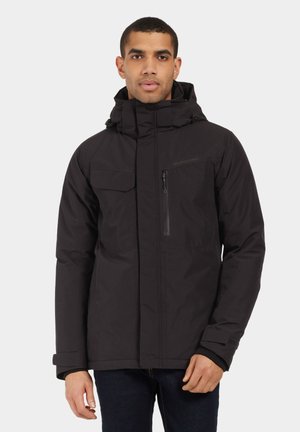 Man wearing a black hooded winter jacket with zip and flap pockets, standing against a plain light background.