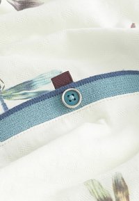 Bouton bleu cousu sur un tissu blanc avec un bord texturé bleu et une petite boucle en tissu marron, partie d'un détail de vêtement avec des impressions discrètes.