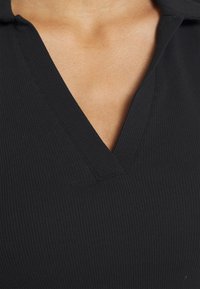 Musta ribbineuloksinen V-kaula-aukkoinen paita, jossa pehmeä kangas. Siinä on taitettu kaulus ja siistit ompeleet kaula-aukon ja helman kohdalla.