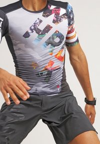 Krátký rukáv sportovní top v šedé barvě s barevnými abstraktními vzory a velkým textem "HRT"; spárován s černými šortkami s matným povrchem.