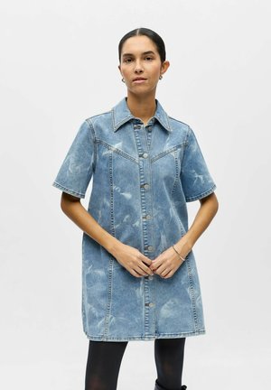 Femme portant une robe en denim délavé bleu clair à manches courtes et boutonnée, avec un legging noir, debout devant un fond clair uni.
