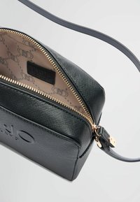 Sac crossbody texturé noir avec fermeture éclair et quincaillerie dorées, présentant un logo et un intérieur beige à motifs. Sangle ajustable comprise.