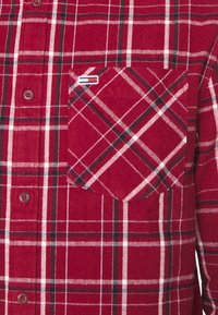 Červená flanelová košile s kostkovaným vzorem obsahujícím bílé a černé pruhy, přední zapínání na knoflíky a náprsní kapsou s logovou nášivkou. Měkká textura a hladký povrch.