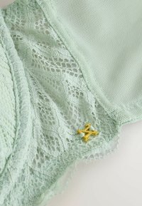 Tissu en dentelle vert clair avec un motif floral délicat, bordure en maille et un petit pendentif doré en forme de "M" attaché près du bord.