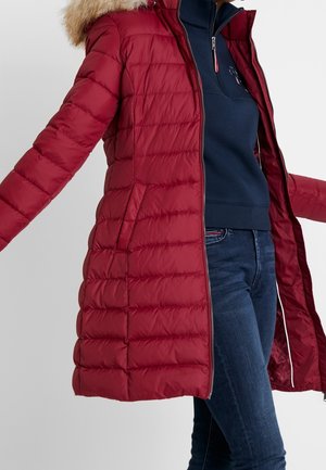 Mujer con un abrigo largo acolchado rojo con capucha bordeada de piel, suéter azul marino con cremallera y vaqueros azul oscuro, con los brazos ligeramente extendidos.