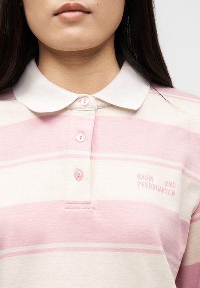 Κοντινό πλάνο ατόμου που φοράει ροζ και λευκή ριγέ μπλούζα πόλο με λευκό γιακά και μικρό λογότυπο με κείμενο στο στήθος.