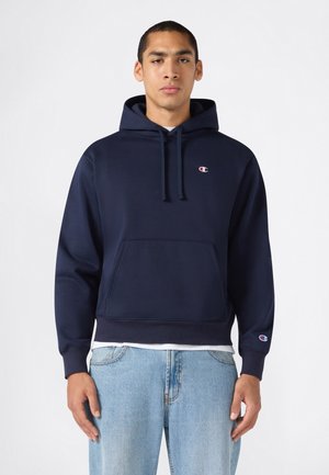 Junger Mann trägt einen marineblauen Champion-Hoodie mit Fronttasche und hellblauen Jeans, steht vor einem schlichten weißen Hintergrund.