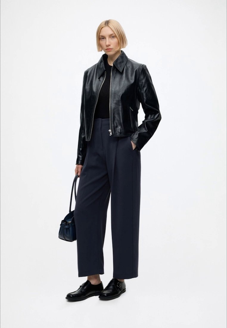 Mujer con cabello corto y rubio, vestida con una chaqueta de cuero negra, top negro, pantalones anchos oscuros, zapatos negros, sosteniendo un pequeño bolso negro.