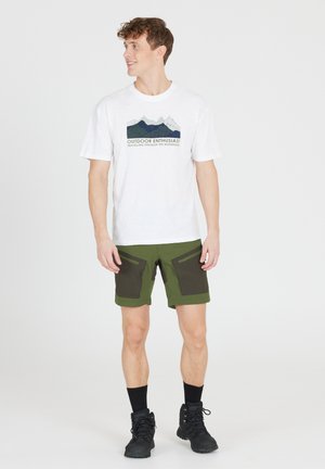 Männermode mit einem weißen Baumwoll-T-Shirt mit Berggrafik und grünen Shorts mit dunklen Taschen, kombiniert mit schwarzen Socken und Stiefeln.