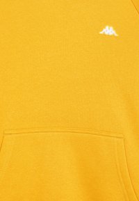 Maglione giallo con una texture morbida, caratterizzato da una tasca frontale e un piccolo logo bianco sul petto, realizzato in un tessuto liscio e misto.