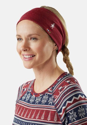 Frau mit blonden Zöpfen, die ein rotes Stirnband und einen rot-weiß-blau gemusterten Pullover mit winterlichen Motiven trägt, lächelt und schaut leicht nach links.