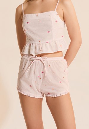 Donna che indossa un completo di pigiama floreale rosa chiaro con canottiera a orlo arricciato e pantaloncini con coulisse su sfondo beige.