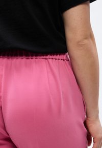 Κοντινό πλάνο ενός ατόμου που φοράει ροζ παντελόνι με ελαστική μέση και μαύρο πουκάμισο, δείχνοντας την πλάτη και το χέρι πάνω σε ουδέτερο φόντο.