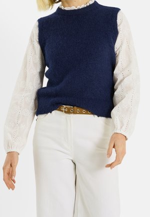 Personne portant un gilet en maille bleu marine par-dessus un chemisier blanc en dentelle à œillets à manches longues, un pantalon blanc et une ceinture marron avec une boucle dorée.