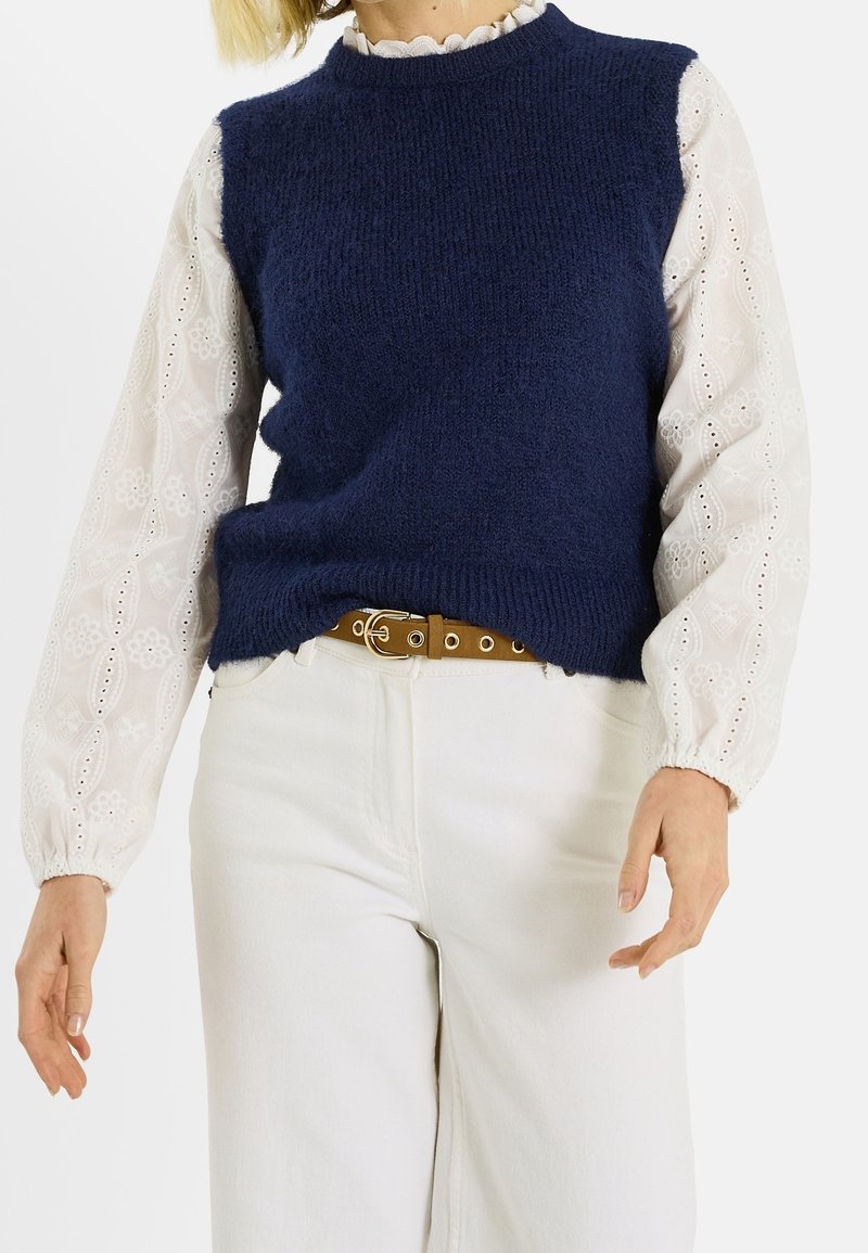 Personne portant un gilet en maille bleu marine par-dessus un chemisier blanc en dentelle à œillets à manches longues, un pantalon blanc et une ceinture marron avec une boucle dorée.