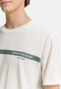 Close-up of a person's neck and shoulder wearing a light beige t-shirt with "DENIM TOM TAILOR EST. 2007" printed in dark green.
