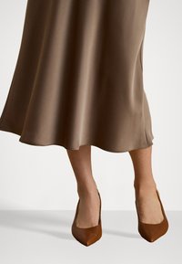 Brun satinklær med en flytende hemline, kombinert med brune spissede hæler. Kjolen har en glatt, skinnende tekstur.