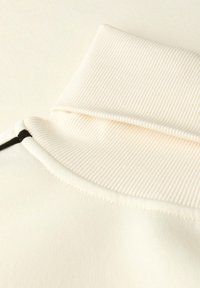 Primo piano di un colletto a dolcevita a costine crema piegato, con un evidente dettaglio di cucitura nera sulla spalla.