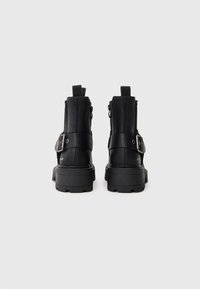 Crne kožne gležnjače s debelim potplatom, zatvaranjem na patentni zatvarač i podesivim remenom s kopčanjem. Glatka tekstura i minimalistički dizajn.