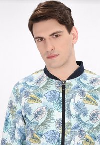 Lichtblauwe zip-up jas met een bladpatroon in groen, geel en blauw, voorzien van een zwarte kraag en ritsdetails.