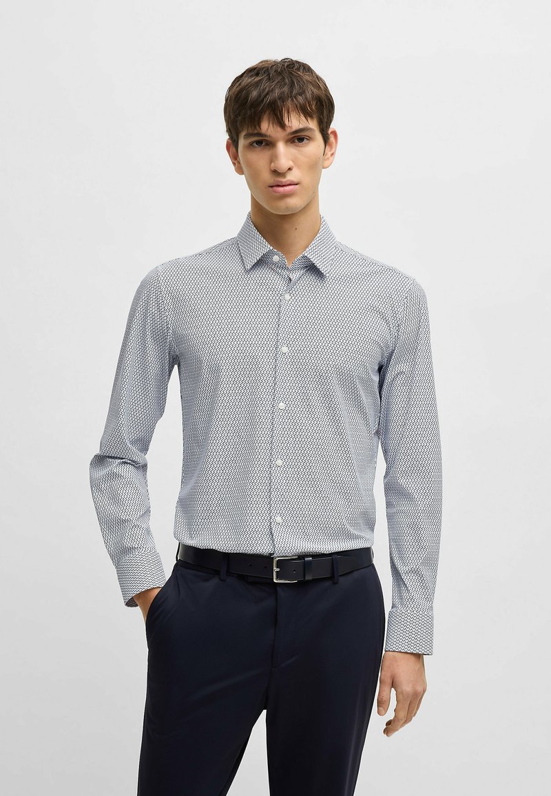 Camicia a maniche lunghe bianca e blu con pattern, colletto button-down, tessuto morbido e design aderente. Abbinata a pantaloni scuri e cintura.
