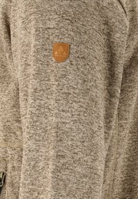 Ljusbrun strukturerad jacka med ett subtilt mönster, med en läderlogotyp. Inkluderar en ficka med dragkedja och mjukt tyg.