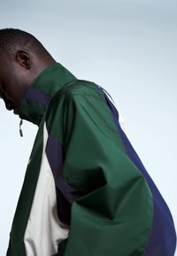 Vihreä, violetti ja beige takki, jossa on korkea kaulus, vetoketjusuljenta ja sileä, kiiltävä pinta. Sivunäkymä esittelee kerroksellisen paneelimuotoilun.