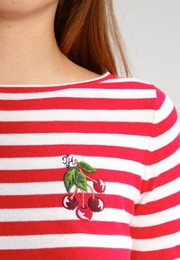 Rød og hvid stribet bluse med bådudskæring, der har et broderet kirsebærdesign og initialen "H" på venstre brystområde.