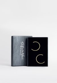 Orecchini a cerchio dorati in una scatola espositiva rivestita di velluto nero con un guscio esterno a texture argentata e logo bianco del marchio.