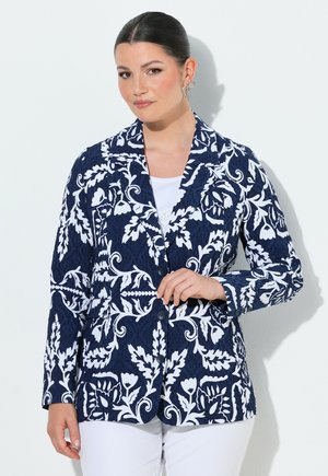 Vrouw draagt marineblauw blazer met witte bloemenpatronen, witte top eronder, witte broek en decoratieve oorbellen, staand tegen een lichte achtergrond.
