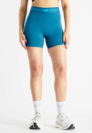 Kvinde iført tætsiddende atletikshorts i teal, hvide sokker og hvide løbesko, stående med hænderne afslappet ved siden.