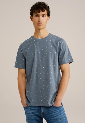 Camiseta azul de manga corta con un patrón geométrico repetido en blanco y acentos metálicos. La tela parece suave y informal.
