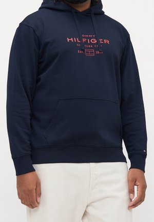 Muž nosící tmavě modrou mikinu s přední kapsou a červeným logem Tommy Hilfiger, v kombinaci s světle béžovými kalhotami.