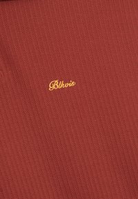 Zbliżenie na czerwonobrunatną, żebrowaną tkaninę z żółtym, haftowanym tekstem w kursywie "Blkvis" wyśrodkowanym na materiale.