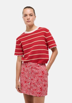 Femme portant un haut en maille rouge et blanc à manches courtes rayées et une jupe rouge boutonnée avec des motifs floraux blancs, debout avec une main dans la poche.