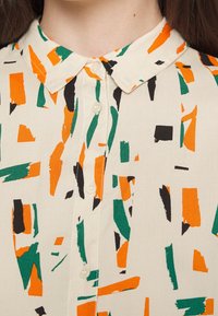 Gros plan d’une chemise crème à col avec des motifs abstraits de coups de pinceau orange, vert et noir et des boutons blancs sur le devant.