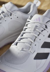 White athletic shoes with mesh texture, featuring black stripes, gray accents, and a rubber sole with purple highlights. Laces are tightly secured.
