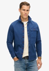 Giacca blu a camicia in denim con colletto, tasca sul petto anteriore e bottoni di legno. Indossata sopra una camicia beige chiaro.