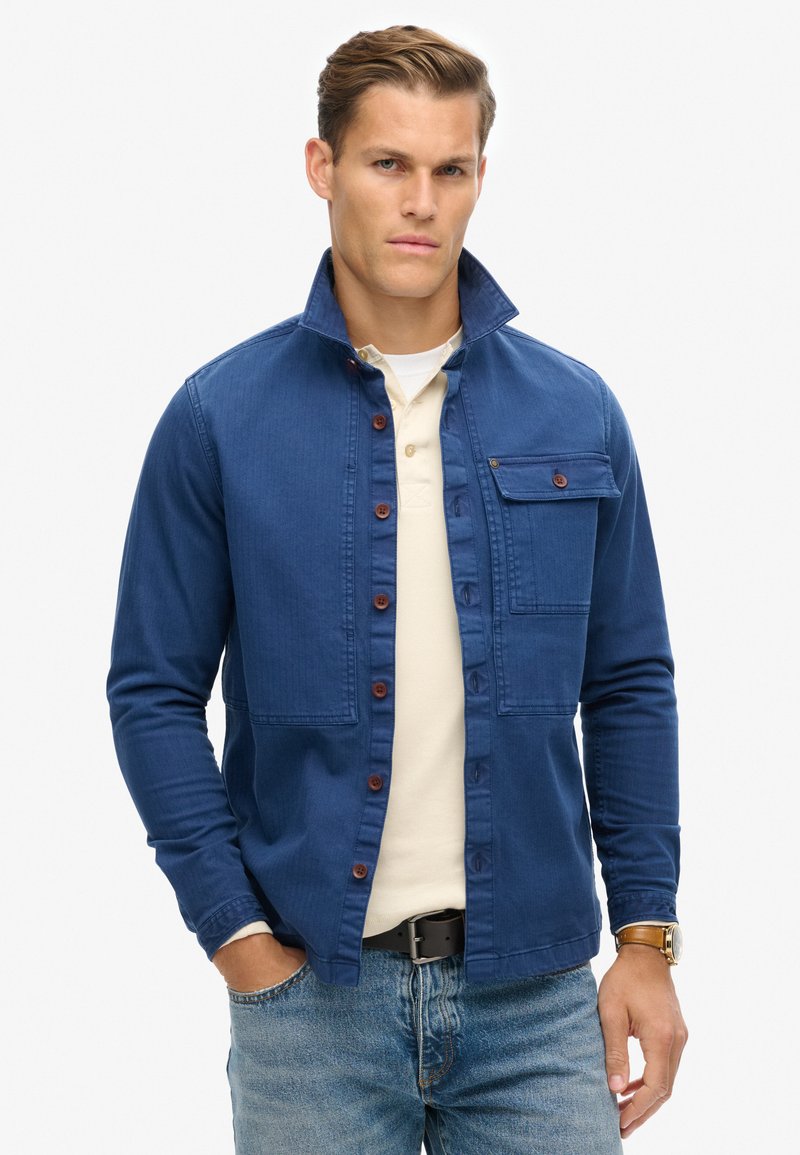 Giacca blu a camicia in denim con colletto, tasca sul petto anteriore e bottoni di legno. Indossata sopra una camicia beige chiaro.