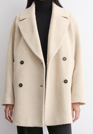 Mujer con un abrigo beige texturizado de doble botonadura sobre un jersey de cuello alto negro, de pie frente a un fondo liso y claro.