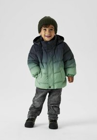 Gepolsterte Jacke in Farbverlauf von Grün zu Grau, ausgestattet mit einem Reißverschluss, Kapuze und Taschen. Kombiniert mit grauen Denim-Hosen und dunklem Schuhwerk.