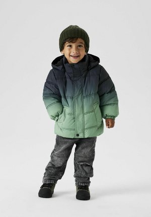 Gepolsterte Jacke in Farbverlauf von Grün zu Grau, ausgestattet mit einem Reißverschluss, Kapuze und Taschen. Kombiniert mit grauen Denim-Hosen und dunklem Schuhwerk.