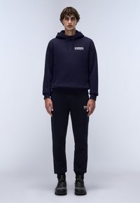 Felpa navy con tasca frontale, polsini aderenti e logo. Abbinata a pantaloni della tuta tapered coordinati e sneakers nere con suole testurizzate.