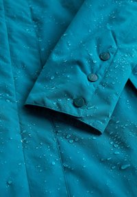 Giacca impermeabile turchese con gocce d'acqua visibili. Presenta un polsino piegato e bottoni con design ancorato, evidenziando una superficie liscia.